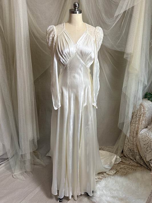 A long white 1930s satin gown with sheer lace sleeves and a gathered bodice, displayed on a mannequin in a bridal-style studio setup with draped tulle and soft fabric backdrop.